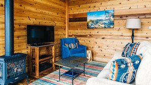 A cozy cabin interior with wooden walls, a television, a blue armchair, a sofa with patterned cushions, and a fireplace.
