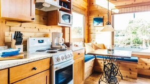 A compact kitchen with wooden cabinets, a microwave, a stove, and a sink. There is a window with a view of greenery outside.