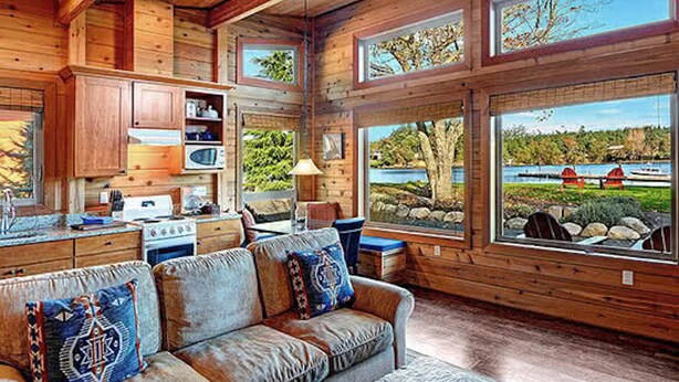 Interior de una cabaña con detalles de madera, un sofá con cojines decorativos, una zona de cocina y grandes ventanales con vistas a un lago.