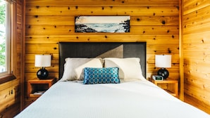 A wooden cabin bedroom with a bed, pillows, a nightstand with a lamp, and a painting of a coastal scene.