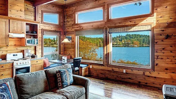 A cabin with wooden walls, a kitchen with a stove, a dining area, and large windows overlooking a lake.