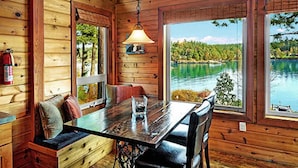 A wooden cabin with a dining table and bench, large windows overlooking a lake, and a hanging lamp.