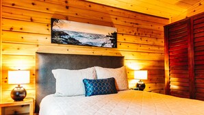 A wooden cabin room with a bed, two nightstands, and a painting of a coastal scene.