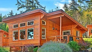 A wooden cabin with a covered porch, large windows, and a garden with various plants. - Snug Harbor Resort and Marina - Two Bedroom Water View Cabin, Queen Beds (Friday Harbor)