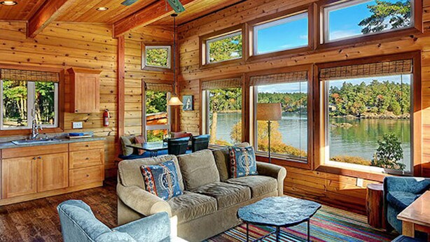 A cabin interior with a wooden ceiling, large windows overlooking a lake, a sofa, and a dining area. - Snug Harbor Resort and Marina - Two Bedroom Water View Cabin, Queen Beds (Friday Harbor)