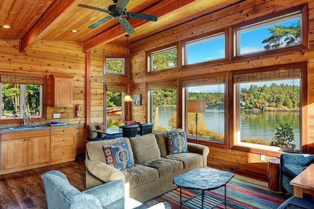 A cabin interior with a wooden ceiling, large windows overlooking a lake, a sofa, and a dining area.