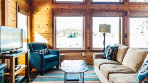 Un interior de cabaña acogedor con paredes de madera, un televisor sobre un mueble de madera, un sillón azul, un sofá beige y una mesita con una lámpara.