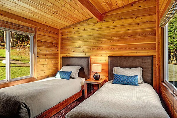 A cabin with two wooden beds, a small table with a lamp, and a window with a view of greenery.