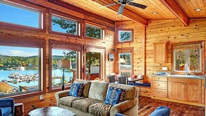 Una cabaña con interior de madera, ventanas amplias que dan a un lago, un ventilador de techo y una zona de cocina.
