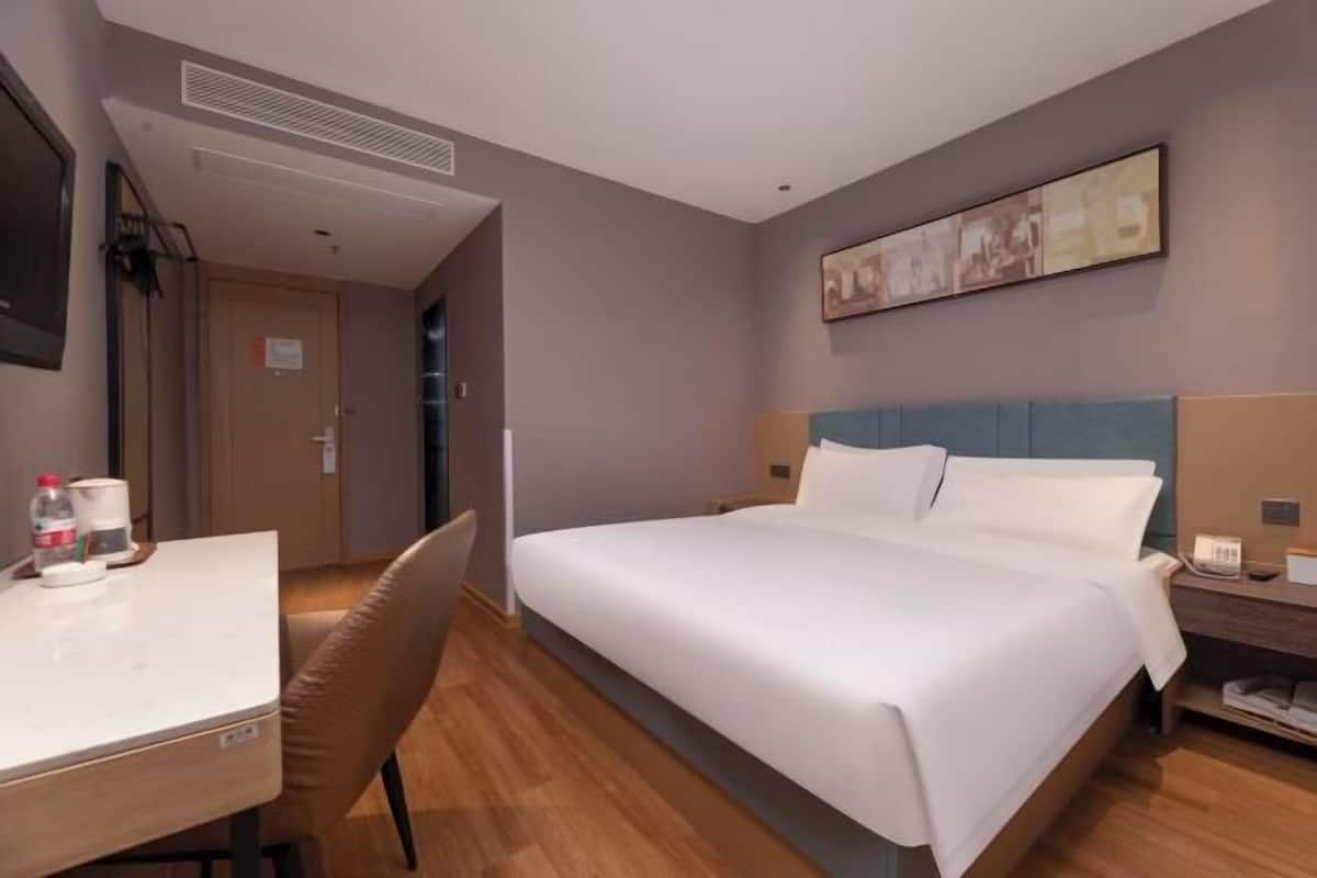 A hotel room with a bed, a desk, a chair, a bedside table, and a wall-mounted artwork.
