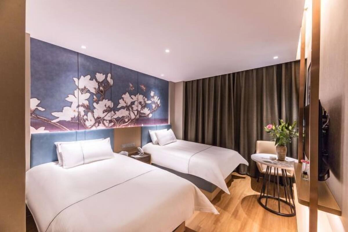 A hotel room with two beds, a small table with a chair, a wall-mounted TV, and a large floral artwork.