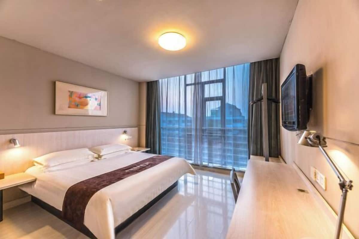 A hotel room with a large bed, bedside lamps, a flat-screen TV, and a window with a view of buildings.