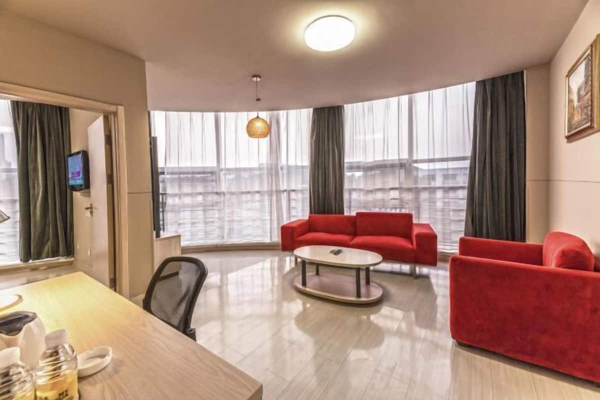 A modern hotel room with a wooden dining table, red sofas, a round coffee table, and large windows with curtains.
