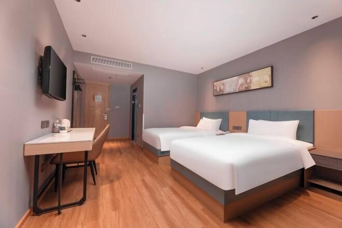 A hotel room with two beds, a desk, a chair, a flat-screen TV mounted on the wall, and a painting above the beds.