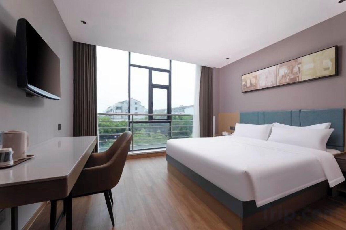 A hotel room with a bed, a desk, a chair, a TV, and a large window with a view.