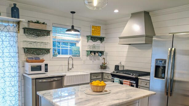 Cozinha moderna com armários brancos, geladeira de aço inoxidável, forno embutido e micro-ondas. Há uma janela com vista para vegetação, e uma prateleira decorativa sobre a pia.