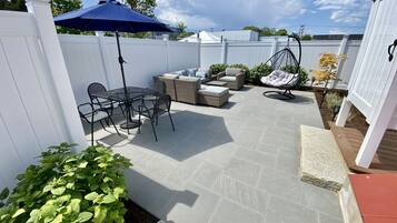 Relax in a serene outdoor space with a glass-top table, cushioned sofa, and hanging chair, surrounded by lush greenery and a shaded area.