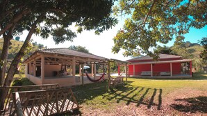 A covered outdoor area with a wooden bench, a hammock, and a red building in the background. - Casa de Campo Rosalinda (Sao Manuel de Carangola)