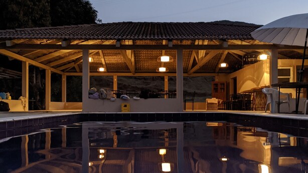 A swimming pool with a covered patio area, a bridge, and a building with a tiled roof in the background. - Casa de Campo Rosalinda (Sao Manuel de Carangola)