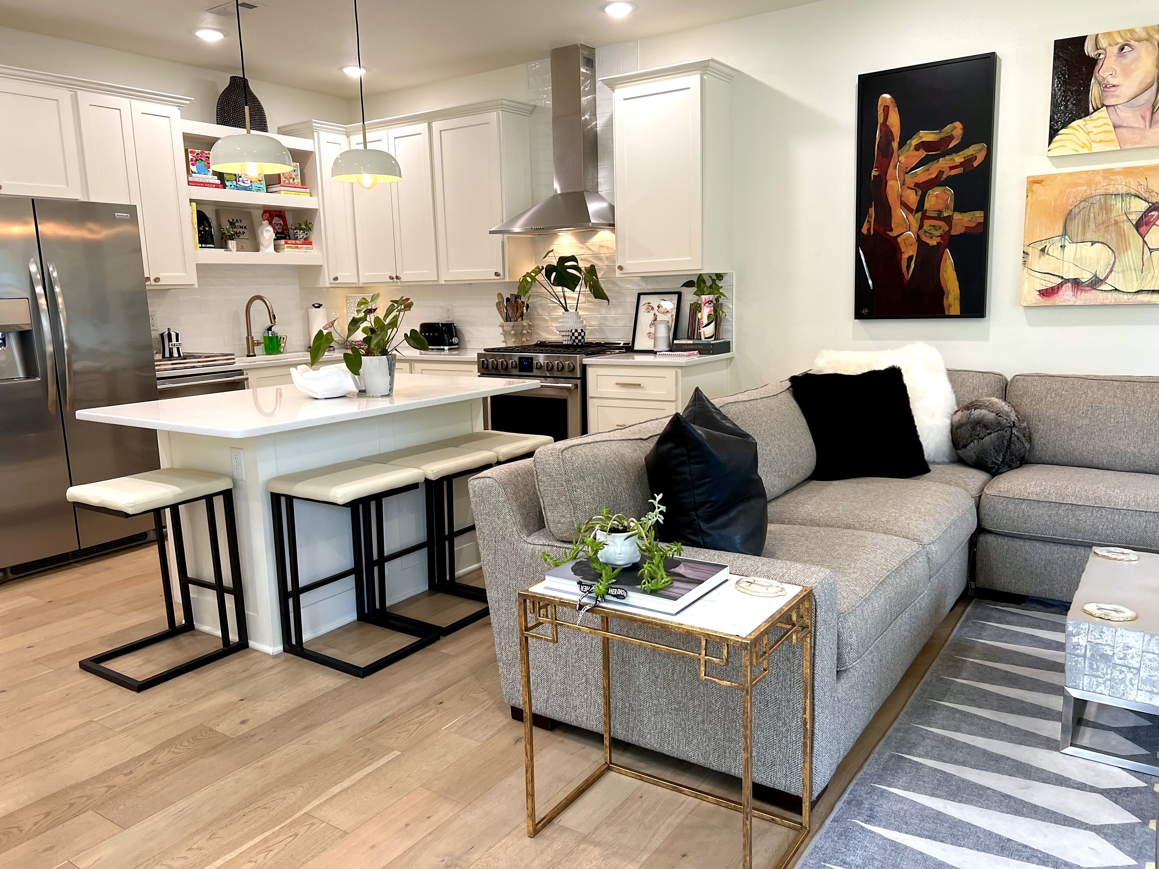 Una cocina moderna con armarios blancos, un refrigerador de acero inoxidable y una isla central con taburetes de bar. Hay un sofá gris con cojines blancos y negros, una pequeña mesa con un florero y un cuadro en la pared.