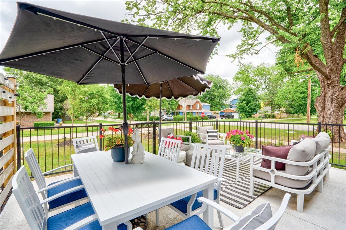 Een terras buiten met een witte tafel en stoelen, een grote parasol en een boom op de achtergrond.