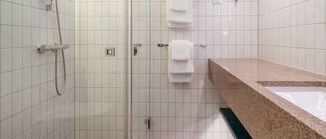 Un baño con paredes de azulejos blancos, una ducha con cabezal de mano, un lavabo con encimera de granito y dos toallas blancas colgadas en un perchero.