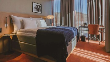 Habitación de hotel con cama, escritorio, silla, lámpara y ventana con cortinas.