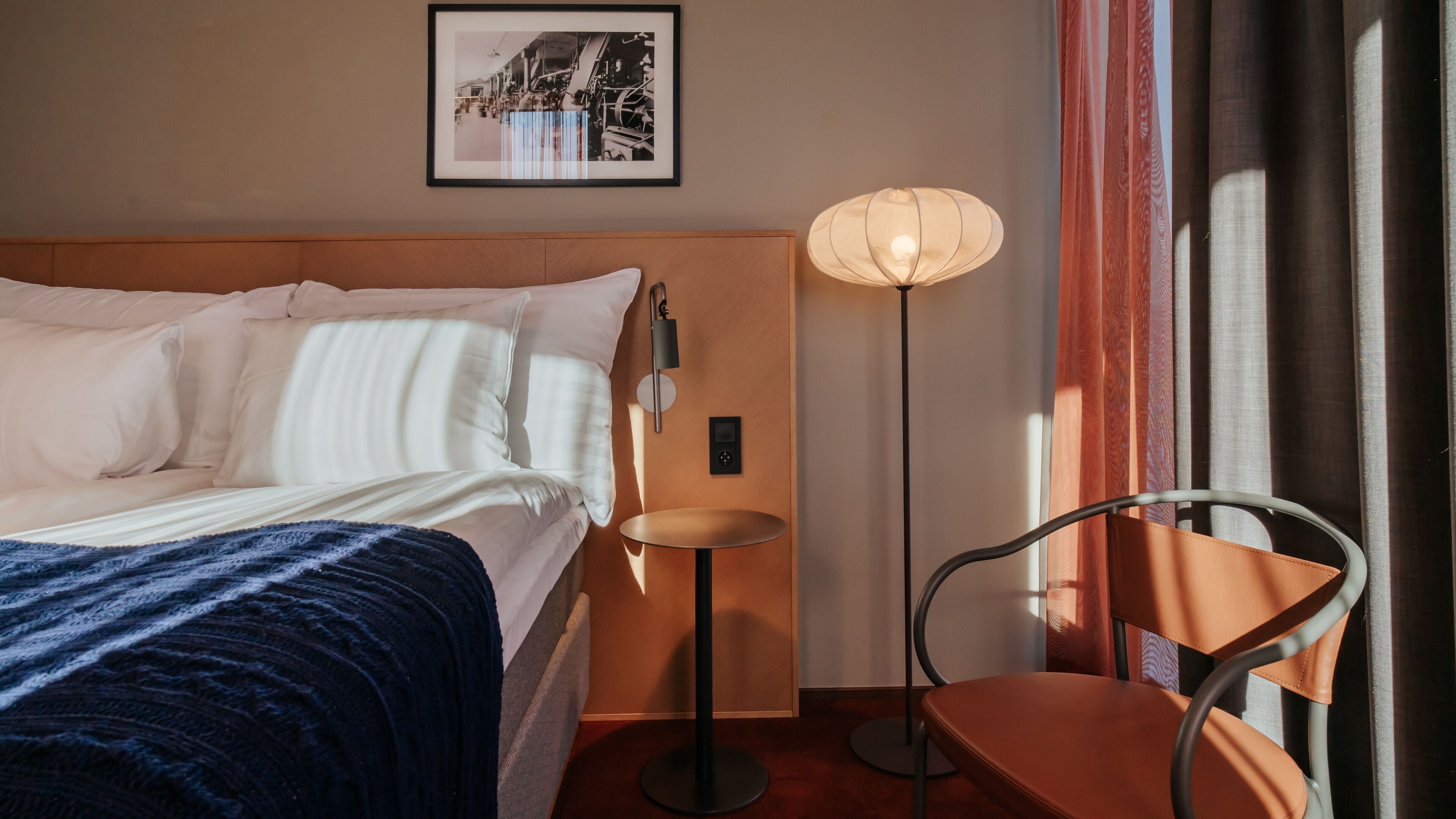 A hotel room with a bed, a small table, a chair, a floor lamp, and a framed picture on the wall.
