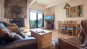 A cozy living room with a stone fireplace, a wooden coffee table, leather sofas, and a dining area with wooden chairs and a table. - CA117 | Lovely Mountainside Condo | Steps from slopes | Winter Shuttle | Hot Tub (Steamboat Springs)