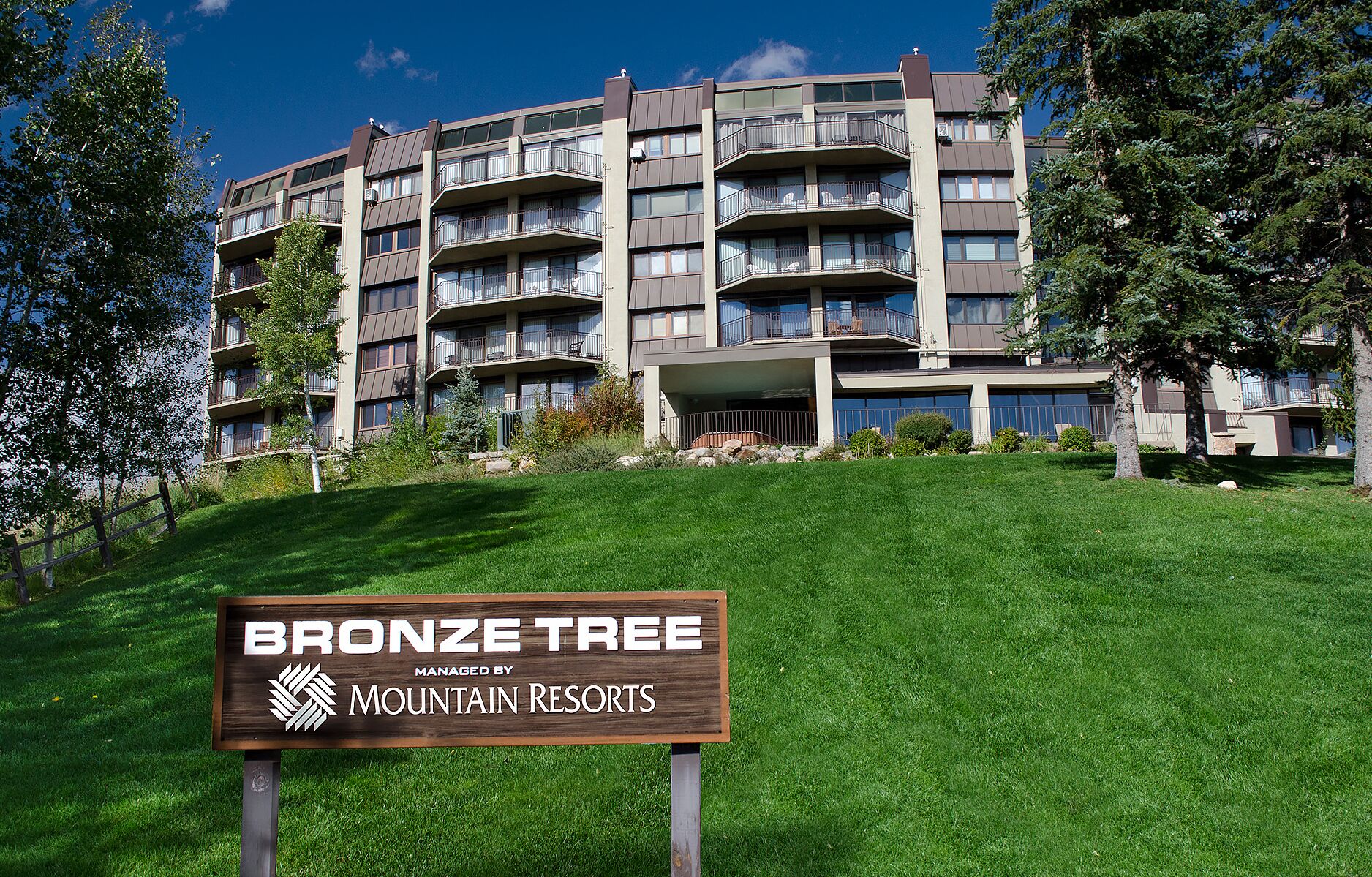 Un immeuble à plusieurs étages doté de balcons, avec un espace verdoyant devant et une enseigne indiquant qu’il est géré par Mountain Resorts.