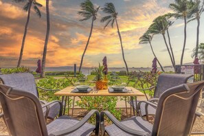 Mesa dispuesta con cuencos, una piña y una botella, con vista a una playa con palmeras y un atardecer.
