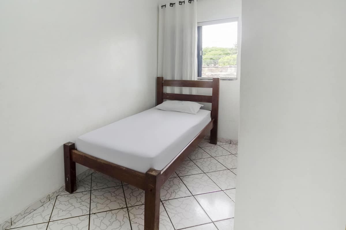Ein Einzelbett mit Holzrahmen und weißer Bettwäsche in einem Zimmer mit weißen Wänden und fußbodenhohen, gemusterten Fliesen.