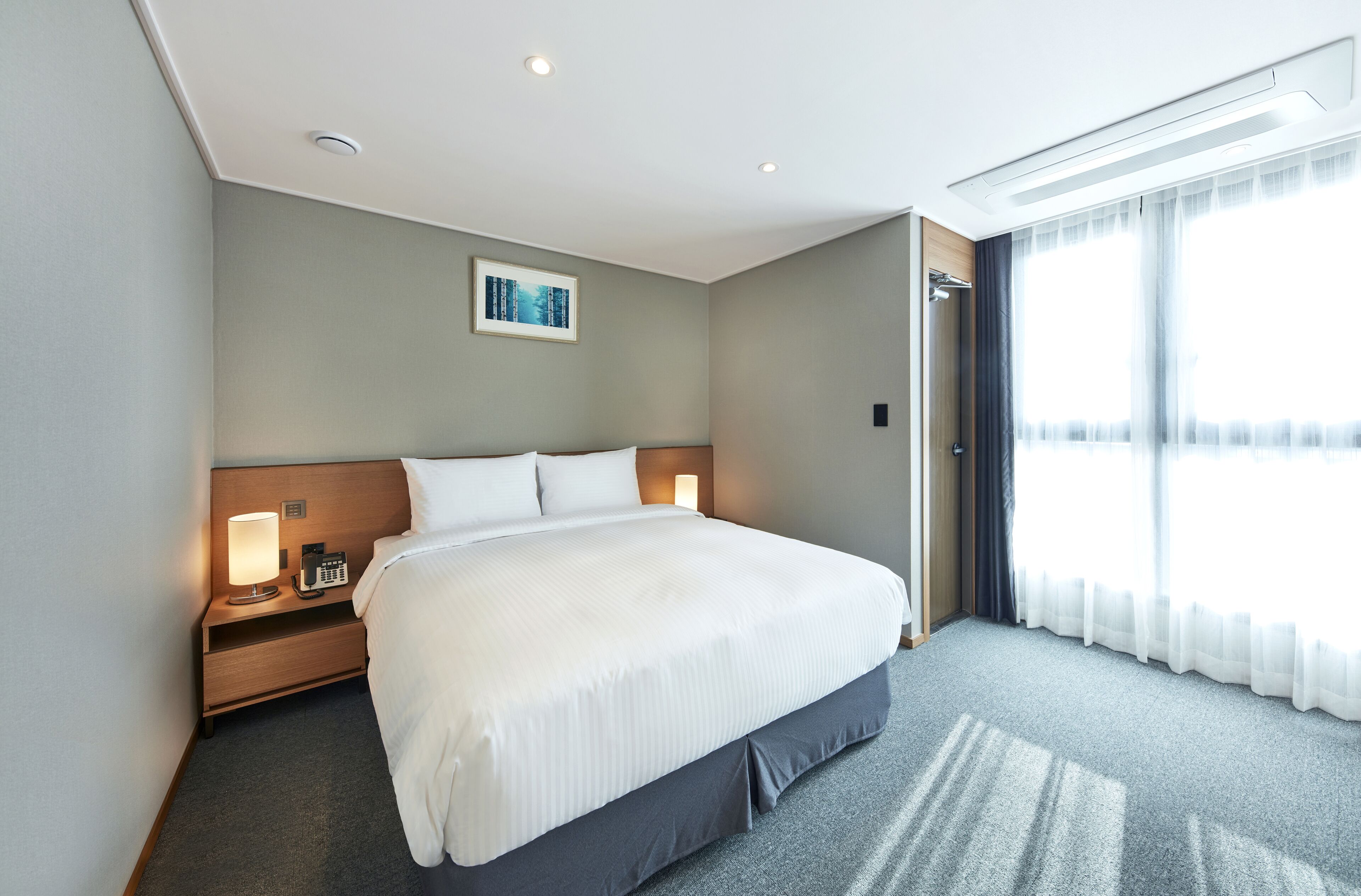 A hotel room with a large bed, bedside lamps, a nightstand, a painting on the wall, and a large window with curtains.