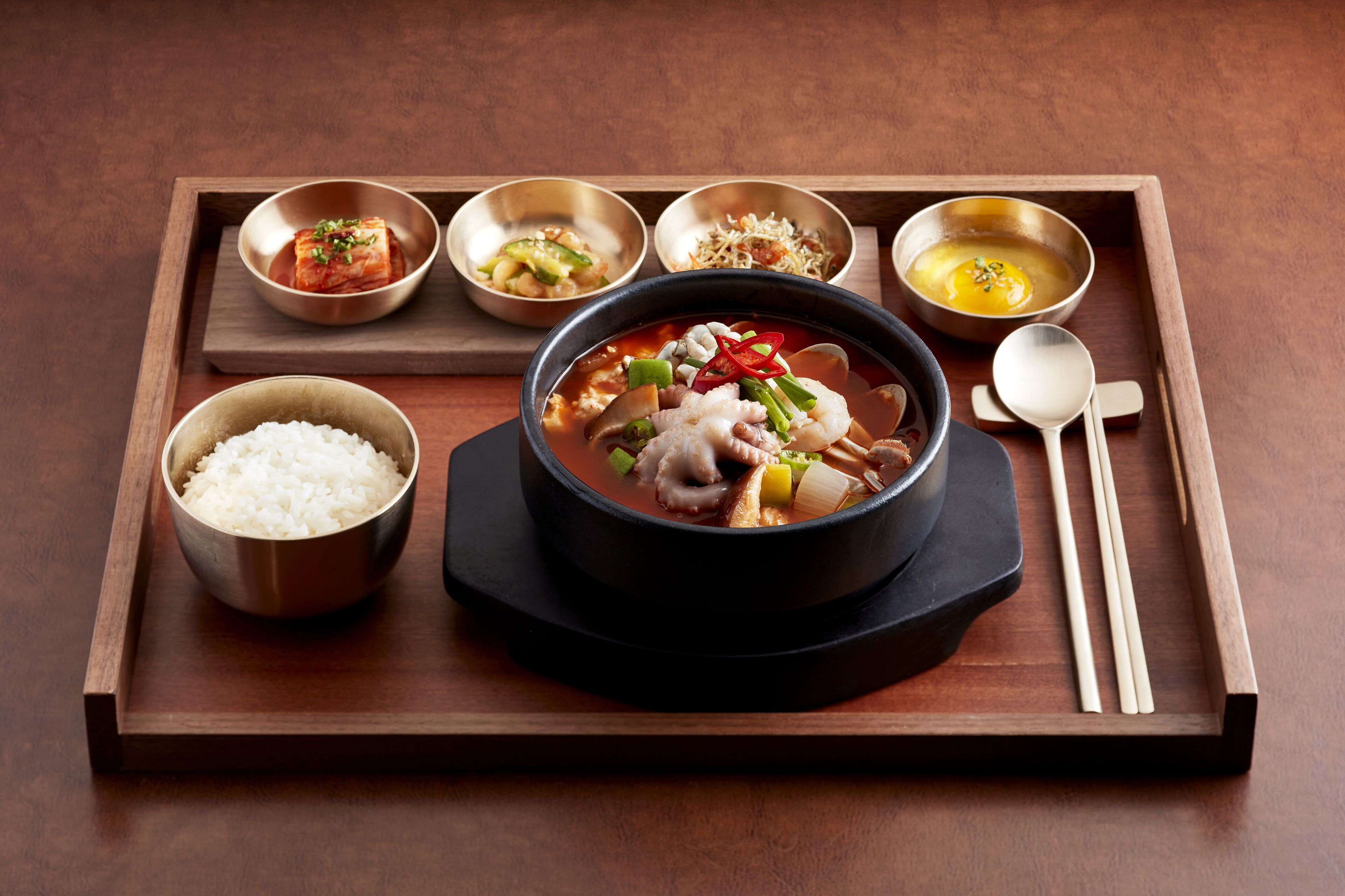 A wooden tray with a bowl of soup, a bowl of rice, and several small bowls containing different side dishes.