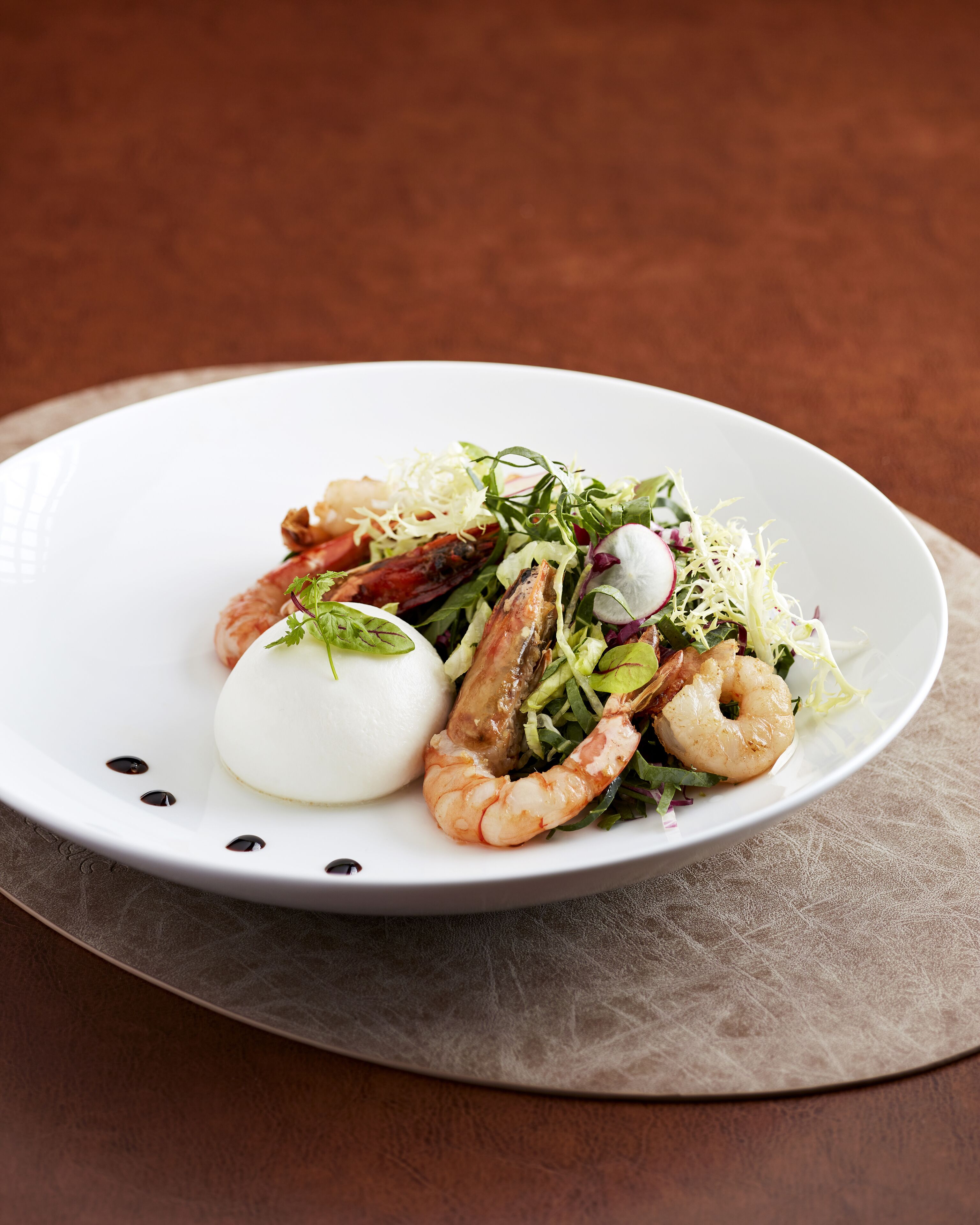 A white plate with a seafood dish, including shrimp and a round white item, possibly a cheese or egg, on a textured surface.