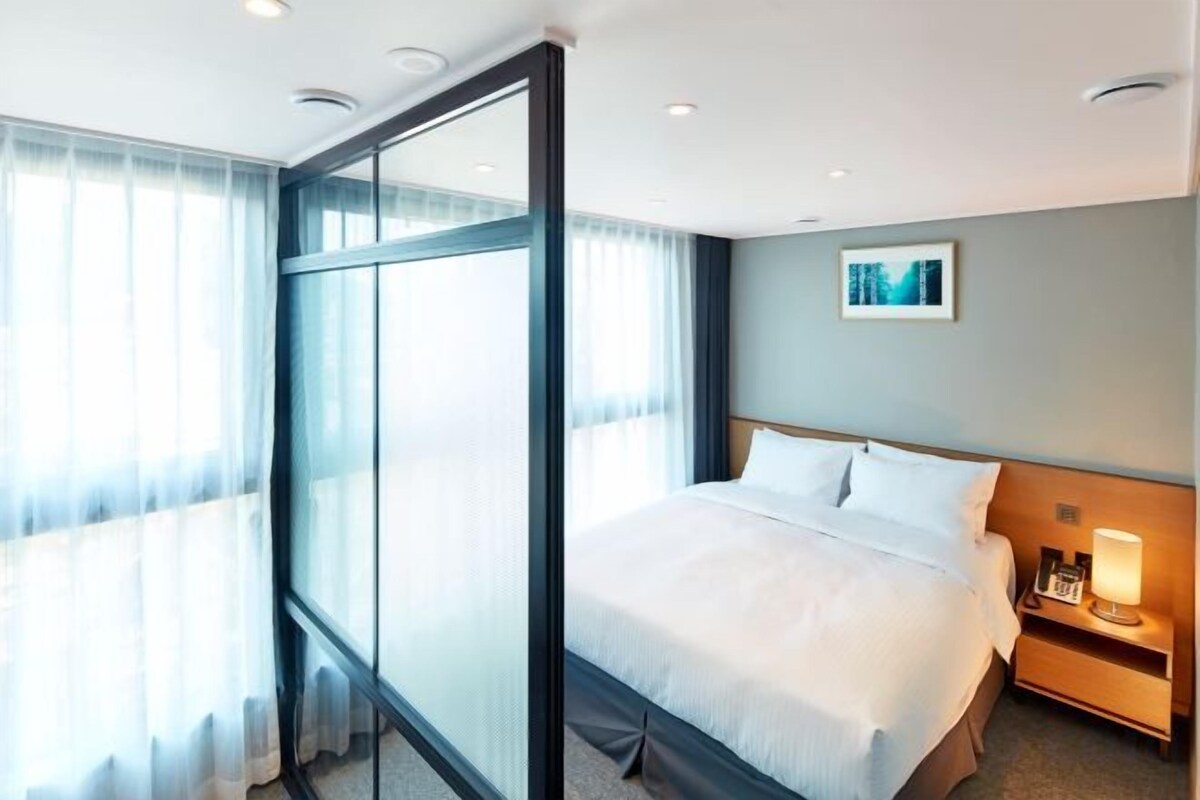 A hotel room with a large bed, a bedside table with a lamp, and a window with sheer curtains.