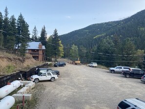 A gravel parking area with several vehicles, including a white SUV and a black pickup truck. There are construction materials and a building with a red roof in the background. - New to market--Cozy Golden Nugget - Efficiency corner unit large balcony  (Red River)