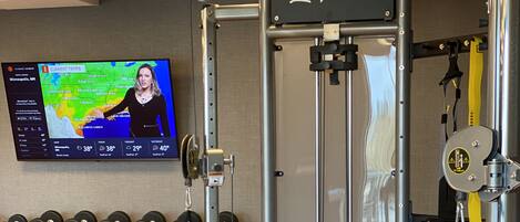 Un gimnasio con una estación de levantamiento de pesas, mancuernas y un televisor que muestra información meteorológica.