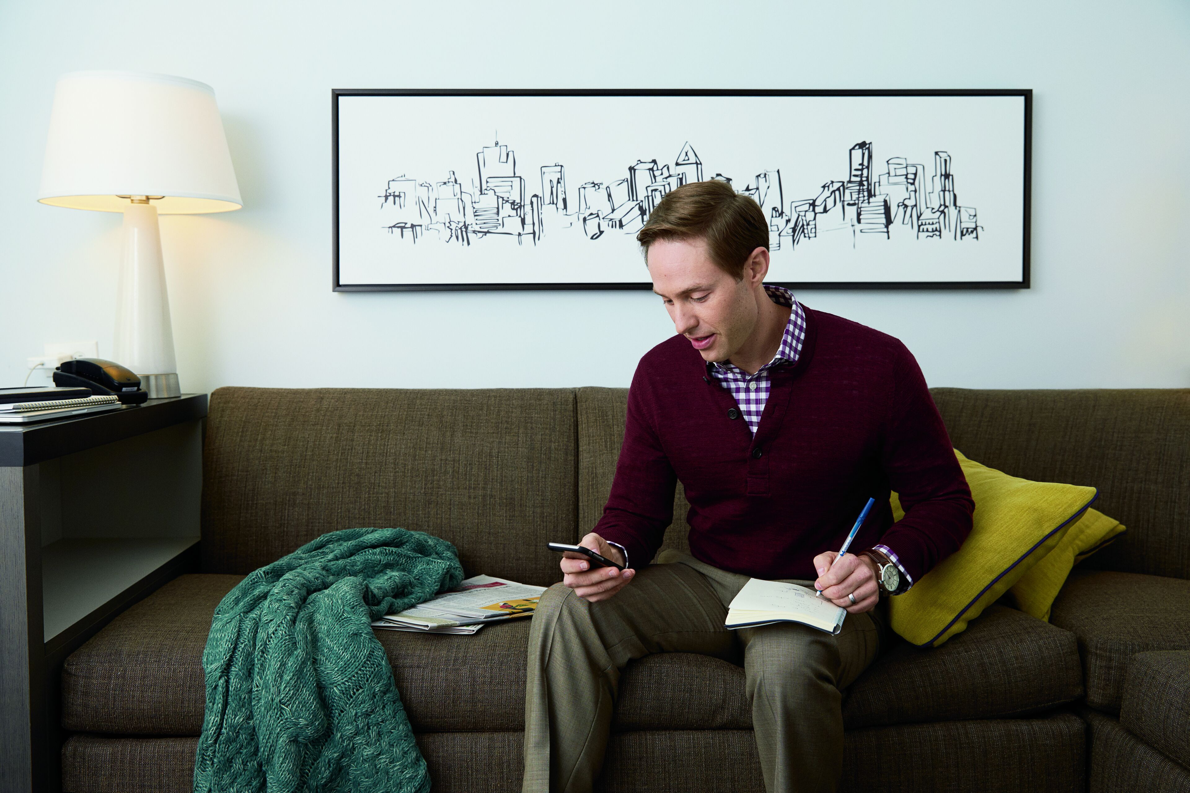 A person sitting on a couch with a lamp, a framed cityscape, and a notebook.