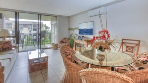 Un amplio espacio habitable con una mesa de comedor de vidrio rodeada de muebles de mimbre, un televisor y vistas al exterior.