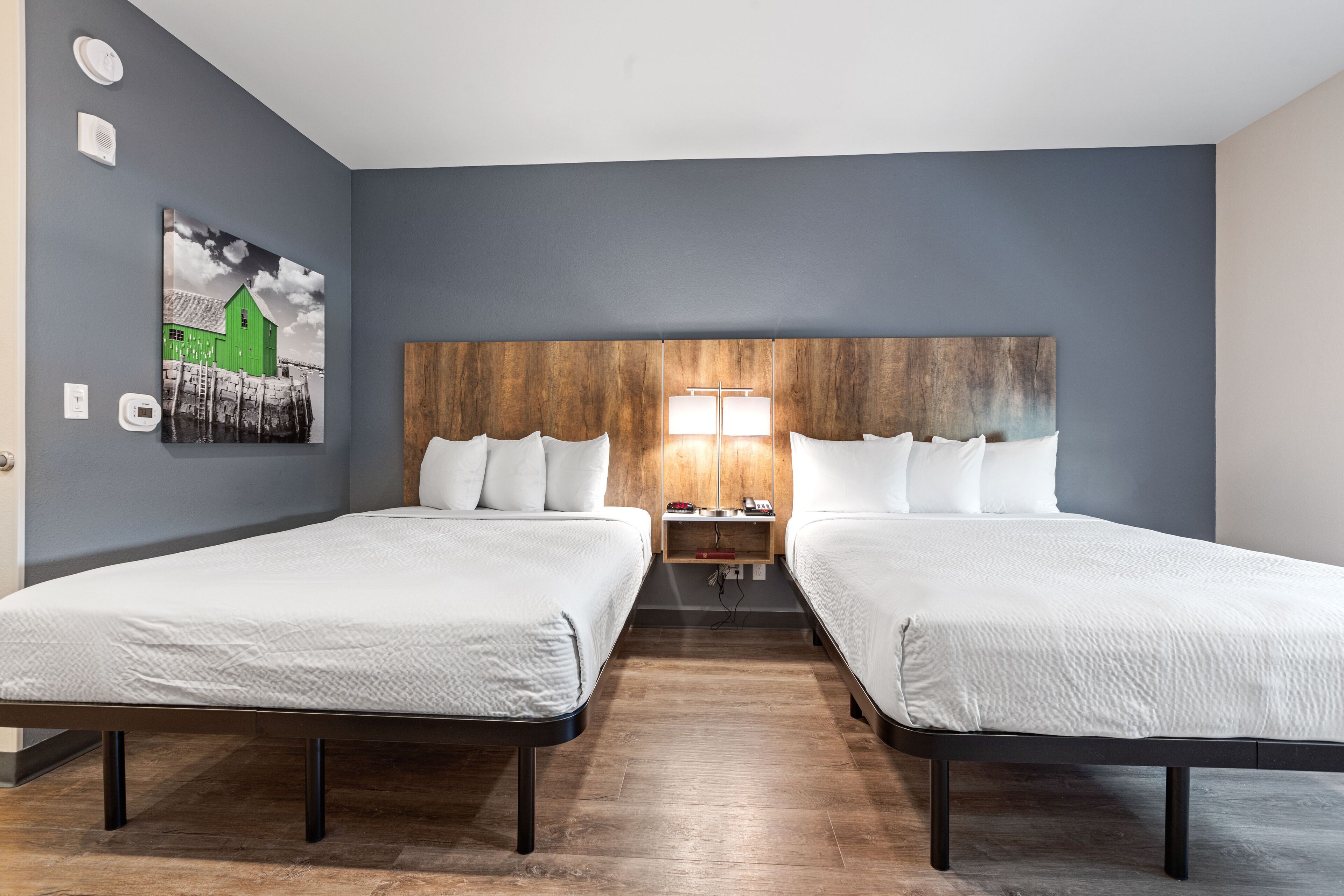 A hotel room with two beds, a wooden headboard, a wall-mounted painting, and a bedside table with a lamp.