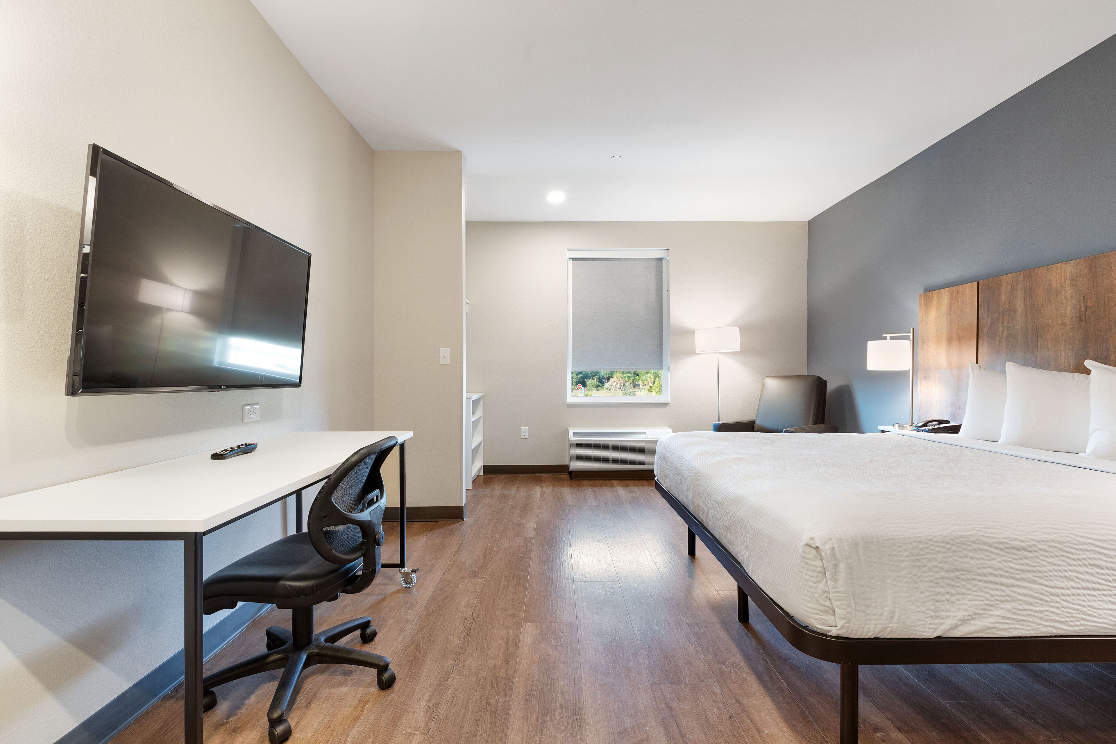 A modern hotel room with a flat-screen TV, a desk, a chair, a bed, a nightstand, a lamp, and a window with a view.