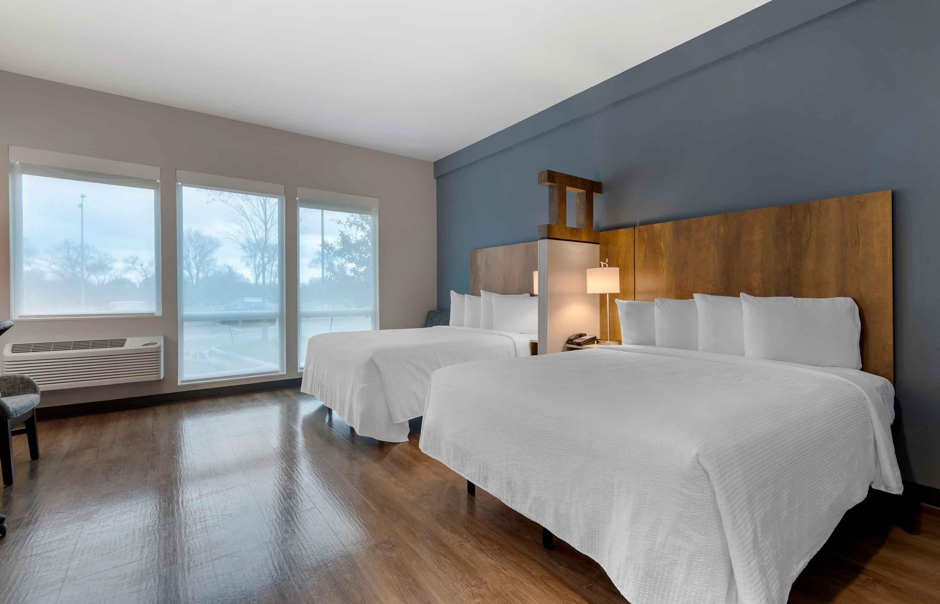 A hotel room with two beds, a large window, a wooden headboard, and a wall-mounted heater.