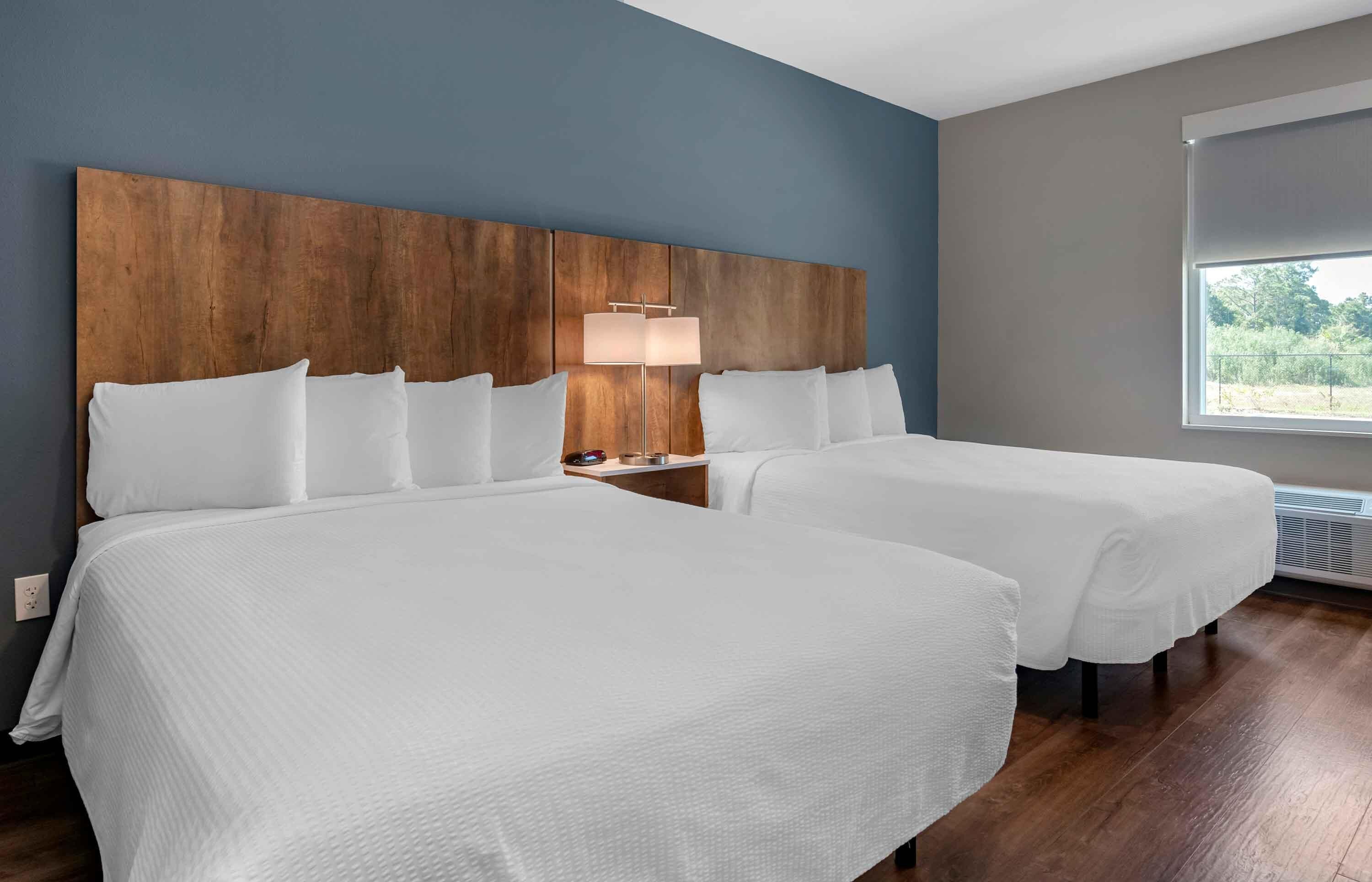 A hotel room with two beds, a wooden headboard, a bedside table with a lamp, and a window with a view.