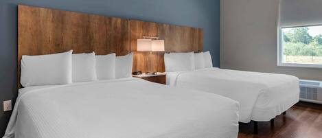 A hotel room with two beds, a wooden headboard, a bedside table with a lamp, and a window with a view.