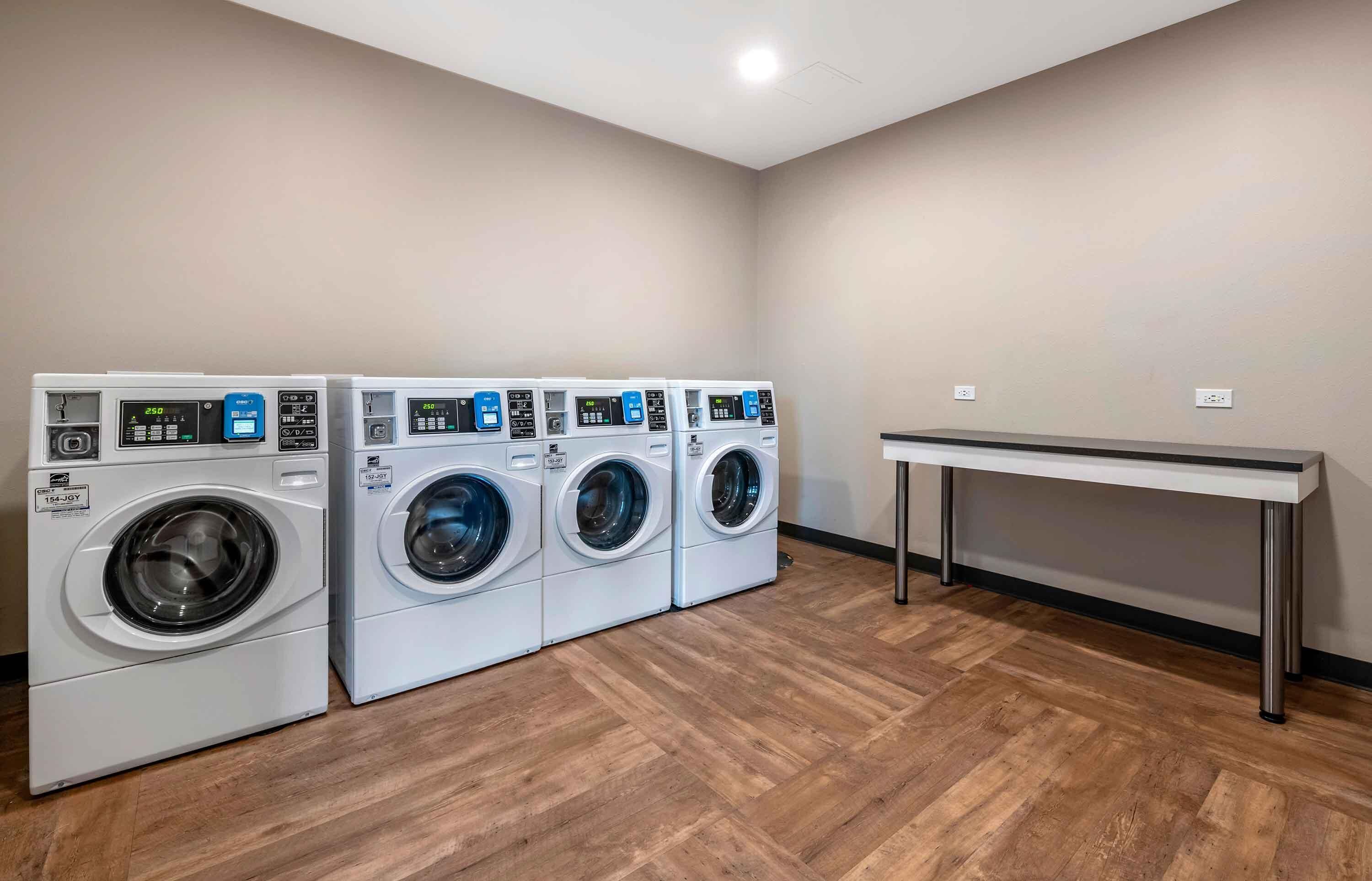 A laundry room with four front-loading washing machines, a table, and a wooden floor.