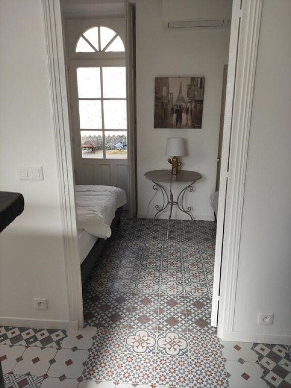 Un couloir étroit avec un lit, une petite table surmontée d’une lampe et des carreaux de sol à motifs.