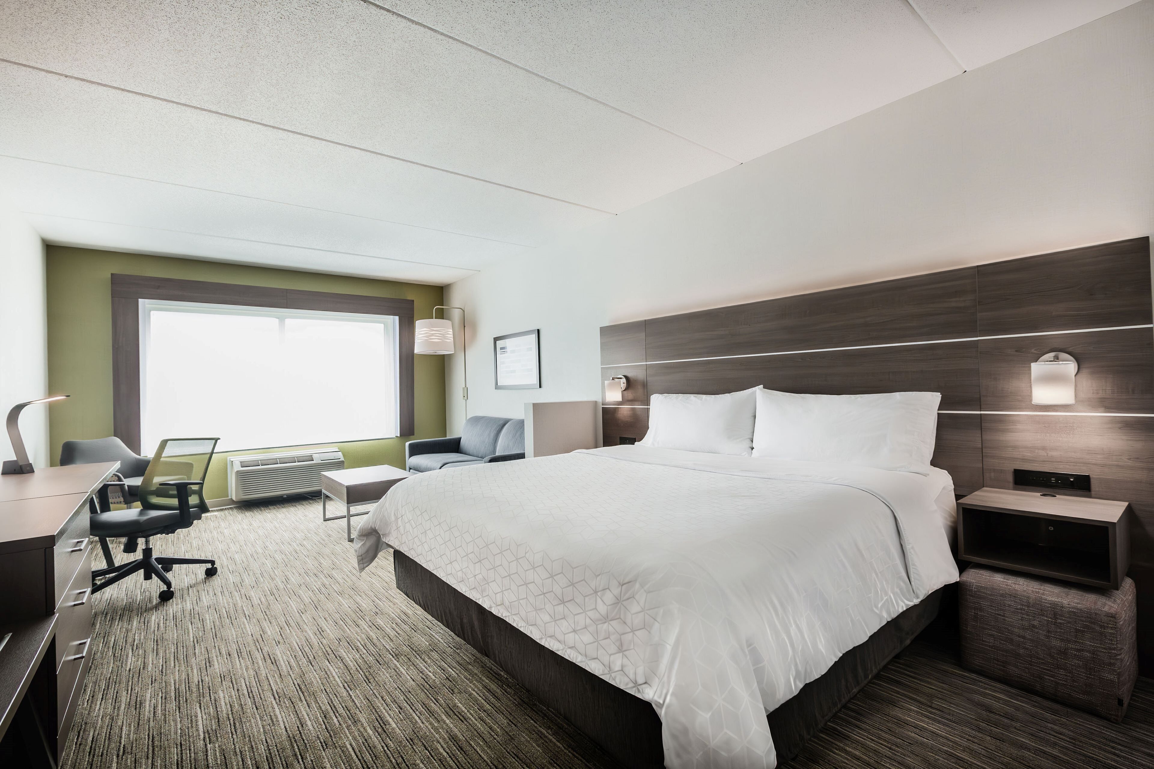 A hotel room with a large bed, a desk with a chair, a small table, and a nightstand with a lamp.