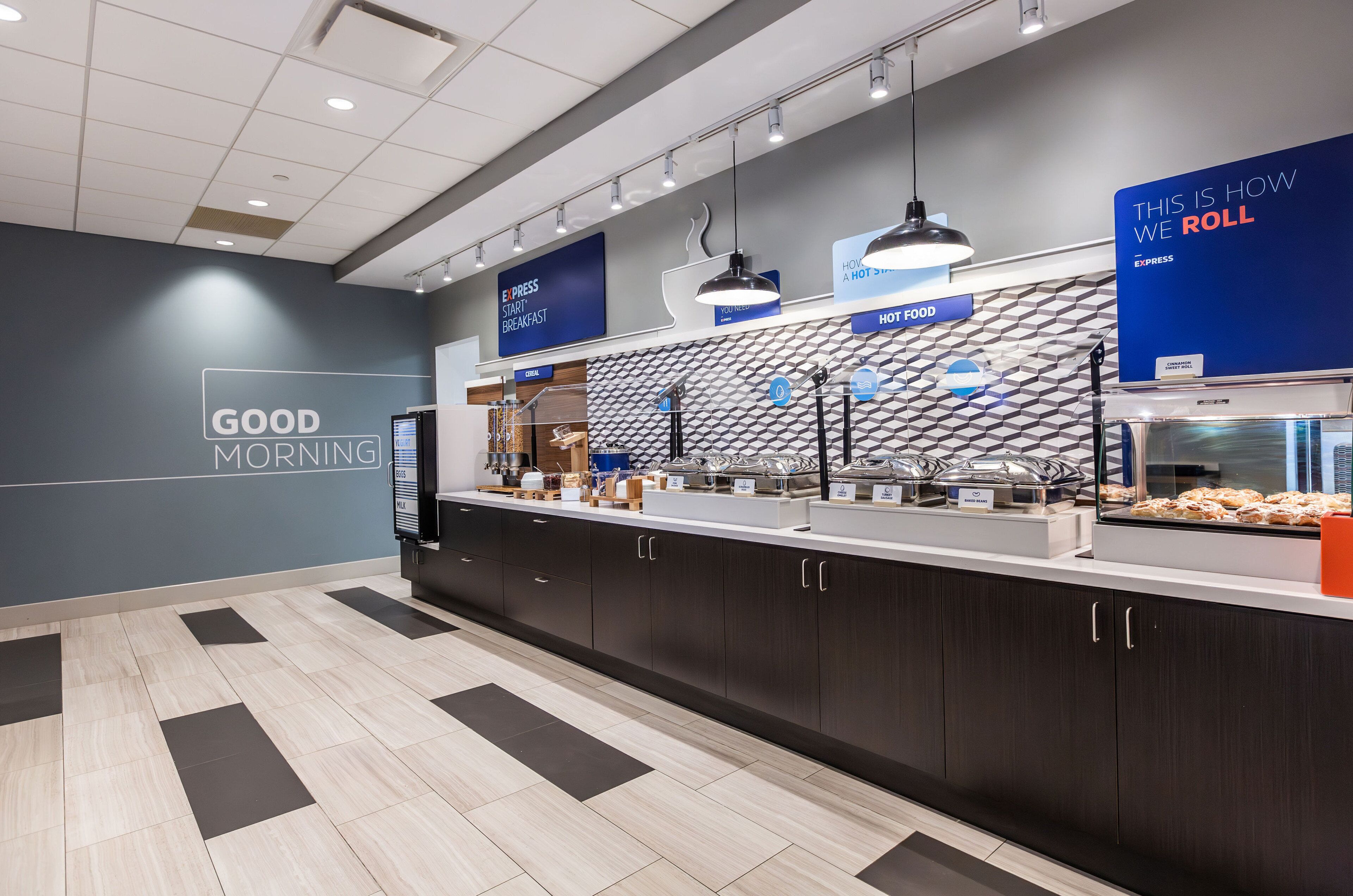 A modern breakfast area with a counter displaying various food items, a sign indicating 'GOOD MORNING', and a wall with a geometric pattern.