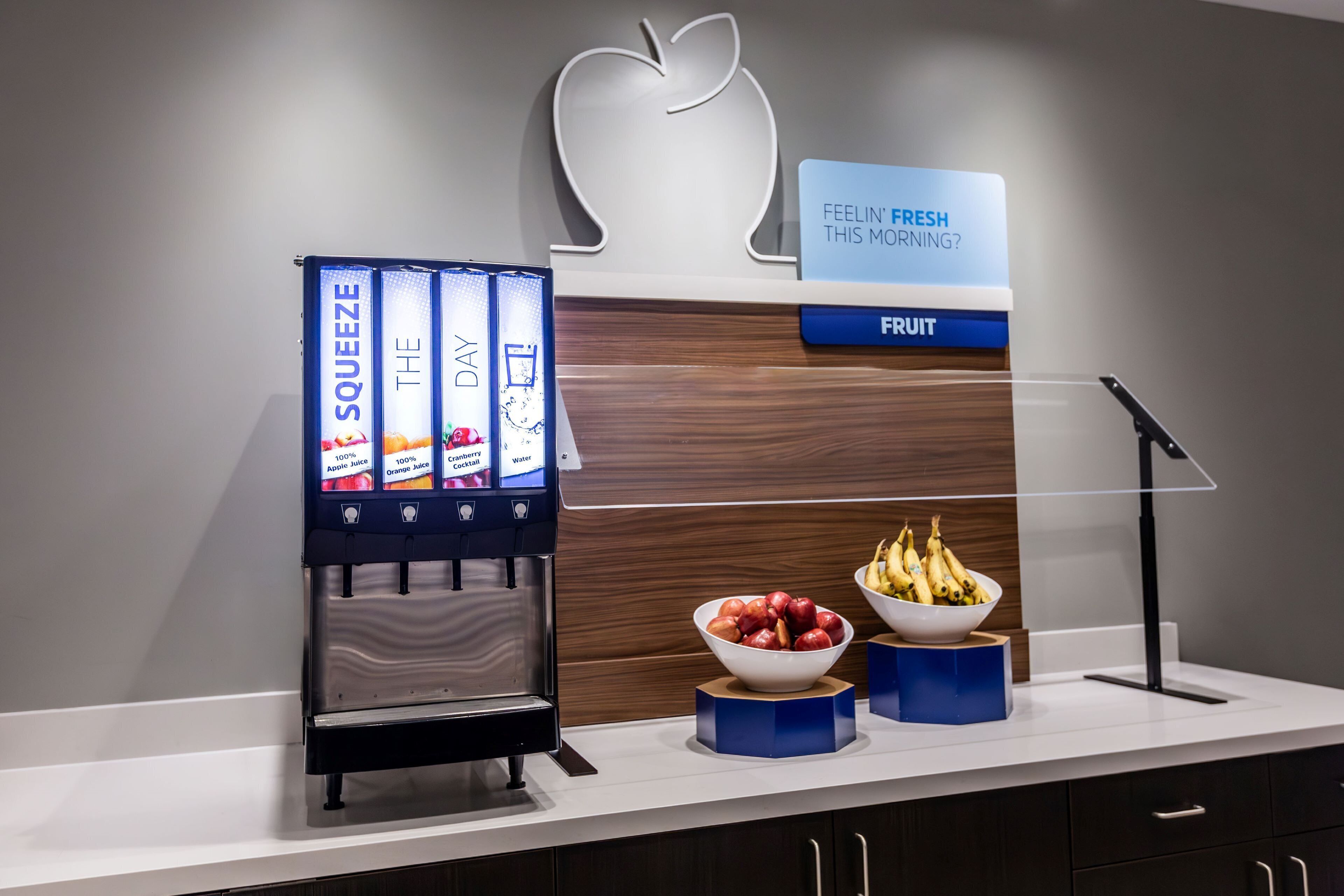 Dispensador de zumos con opciones de manzana, naranja, arándano y agua. Hay cuencos con manzanas y plátanos en la encimera.